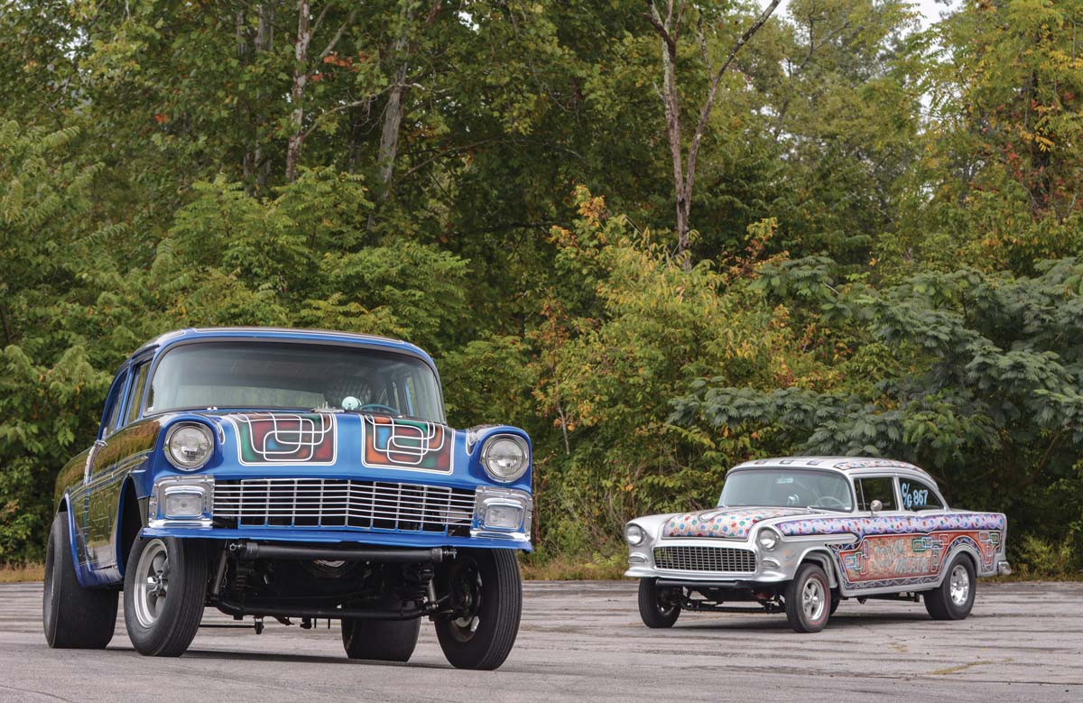 Two brightly-painted Chevrolet Tri-Five Gasser builds on a road in front of a copse of trees