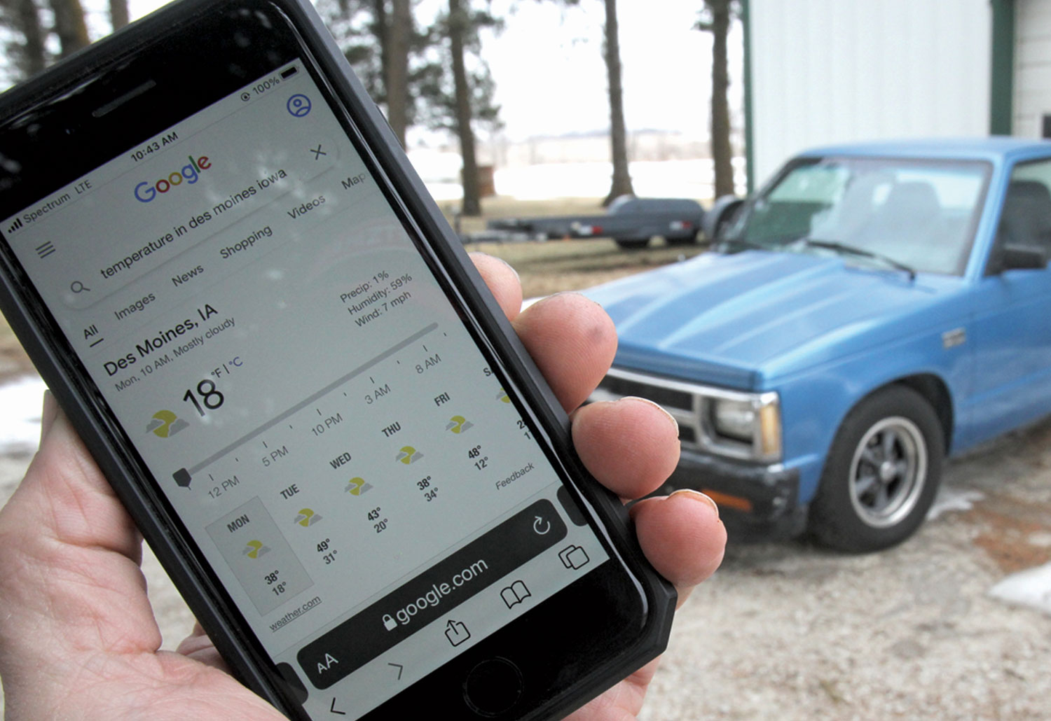 mechanic holds a smartphone showing the temperature of Des Moines, IA while the S-10 truck sits parked in the background