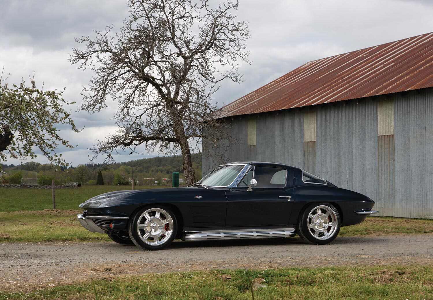 driver's side profile view of the '63 Corvette Sting Ray parked on a dirt road near a large shed