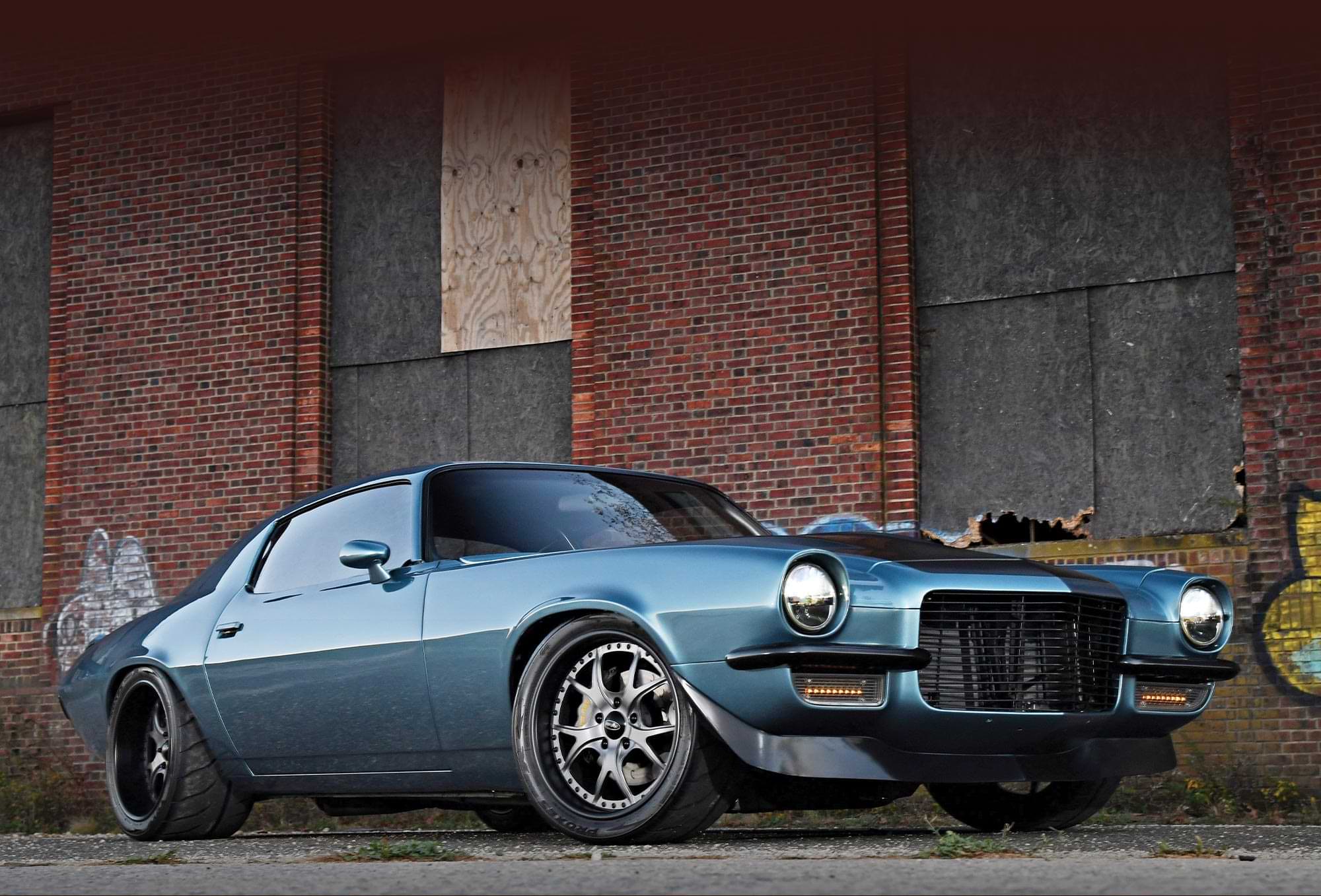low angle three quarter view of Anthony Cacioppo's slate blue and black '71 Camaro parked beside a graffitied brick wall