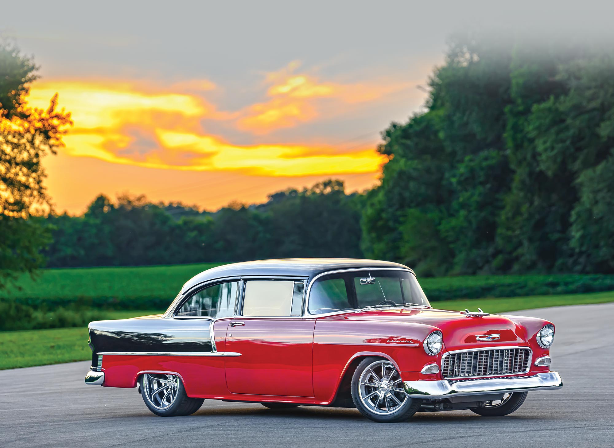 three quarter passenger side view of Darrin Gartrell's black and red '55 Chevy 210 parked against a bright sunset behind deep green trees