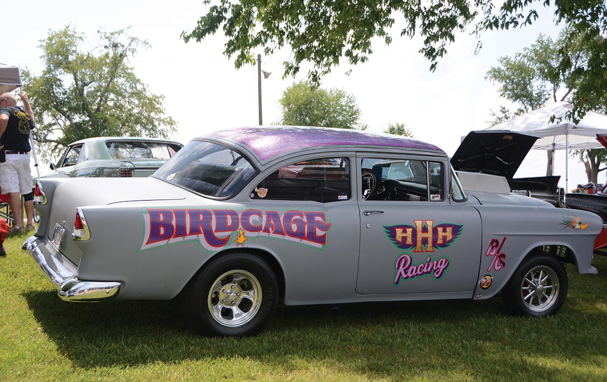 Landscape daytime side photograph perspective of a grey/purple vintage car (named "Bird Cage") on display in a grassy area underneath a tree providing some shade at Wally Parks Nostalgia Nationals in Beech Bend Raceway Park (Bowling Green, Kentucky)