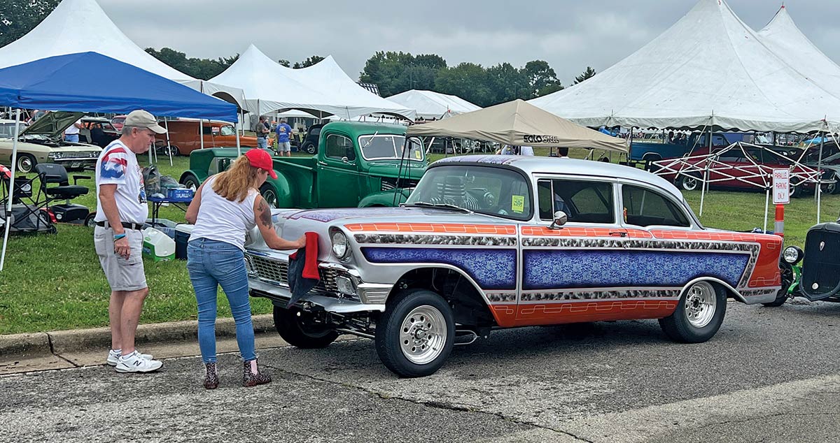 Close-up photograph perspective of a grey/orange/blue vintage Chevy car parked on the curb nearby a grass field area with other tents close by at the 54th annual NSRA Street Rod Nationals as there are two people examining the vehicle