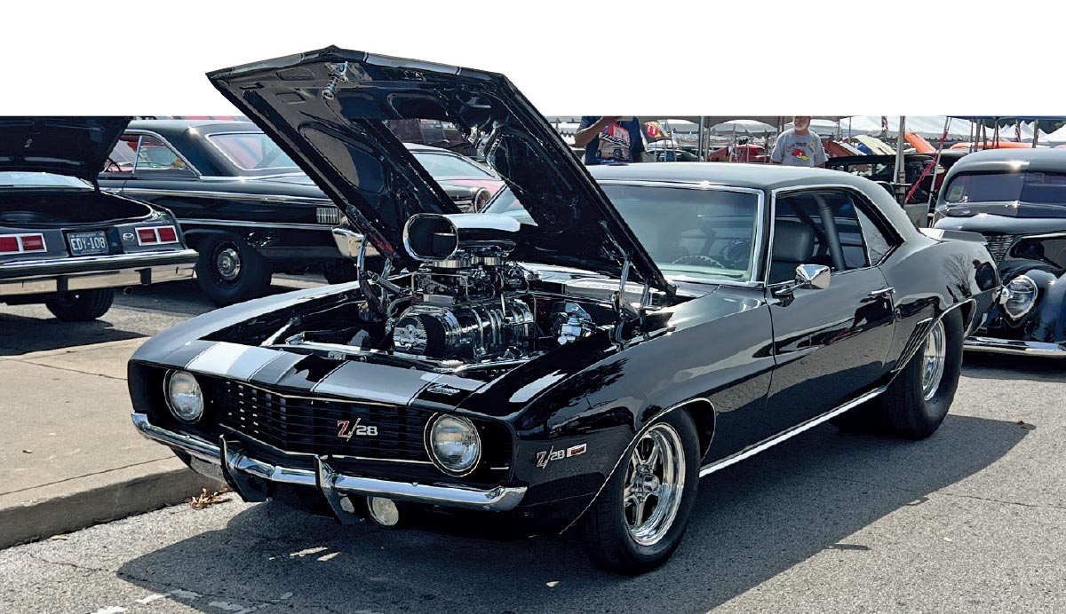 Close-up photograph perspective of a shiny black Chevy vintage car parked on display with its hood opened up at the 54th annual NSRA Street Rod Nationals nearby a curb and other cars in the background