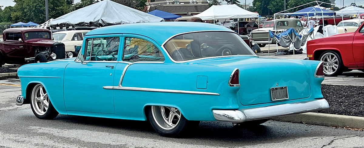 Close-up photograph perspective of a light sky blue Chevy vintage car parked on display at the 54th annual NSRA Street Rod Nationals nearby a curb with other cars and tents around