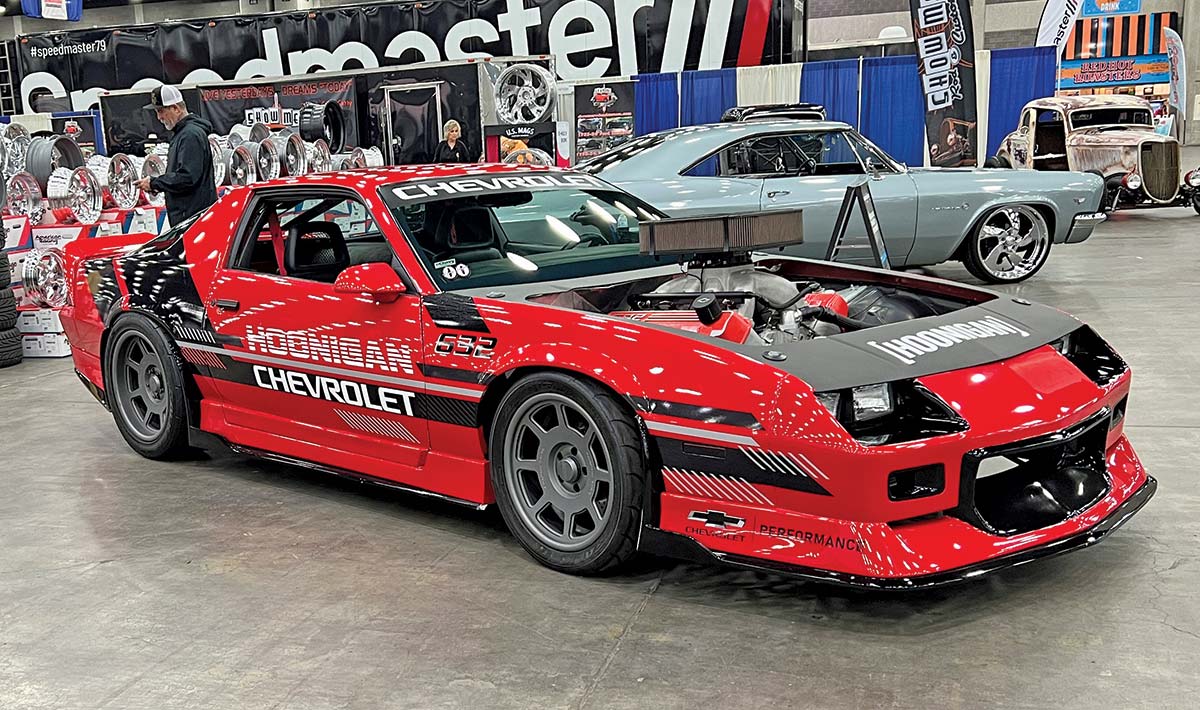 Close-up photograph perspective of a shiny red/black/white Chevy vintage race performance car "Hoonigan" parked on display with the top front hood exposed at the 54th annual NSRA Street Rod Nationals with other cars nearby and two individuals in the area