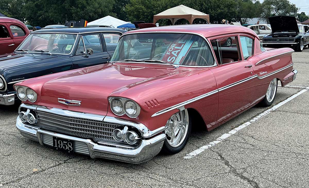 Close-up photograph perspective of a shiny dark pink Chevy vintage car parked on display at the 54th annual NSRA Street Rod Nationals with other cars nearby in the area