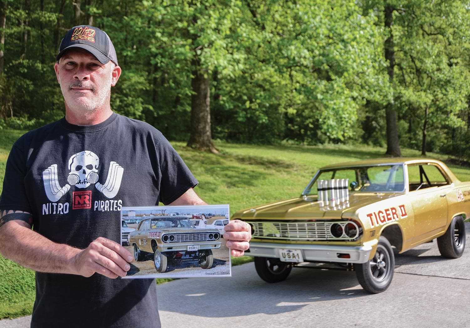 Dave Giles stands holding an image of the gold ’65 Chevelle 300, parked among other performance cars, while standing in front of the gold ’65 Chevelle 300