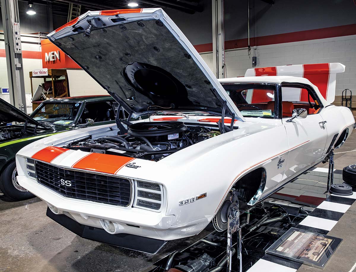 Close-up photograph perspective of a vintage white/orange Camaro muscle car (SS 396 model) with its hood and trunk open plus no tires installed in a display area of a showroom floor