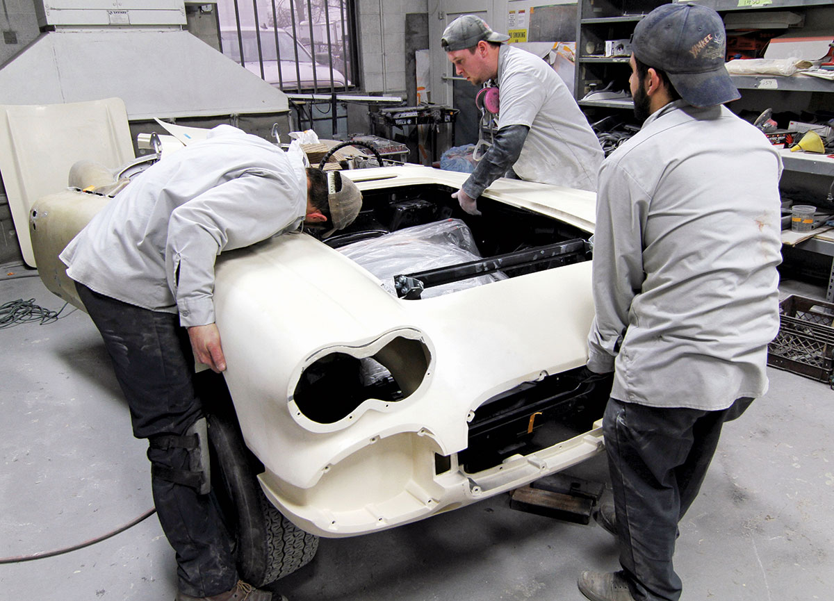 Three men in gray shirts and pants working on a white body of a car being restored