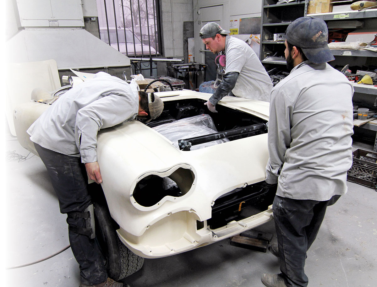 Three men in gray shirts and pants working on a white body of a car being restored