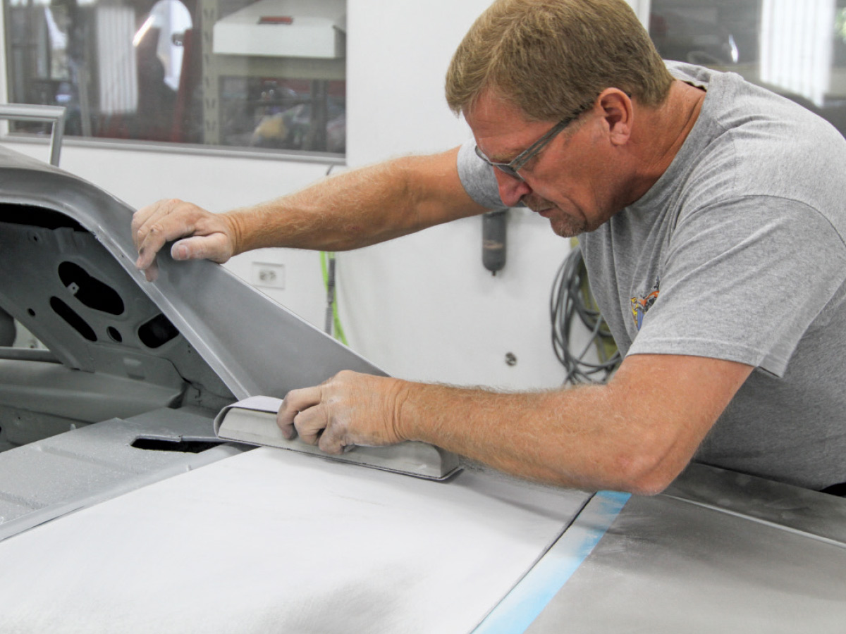 Man wearing grey t-shirt and eye protection using 80-grit sandpaper to even out filler on sheet metal