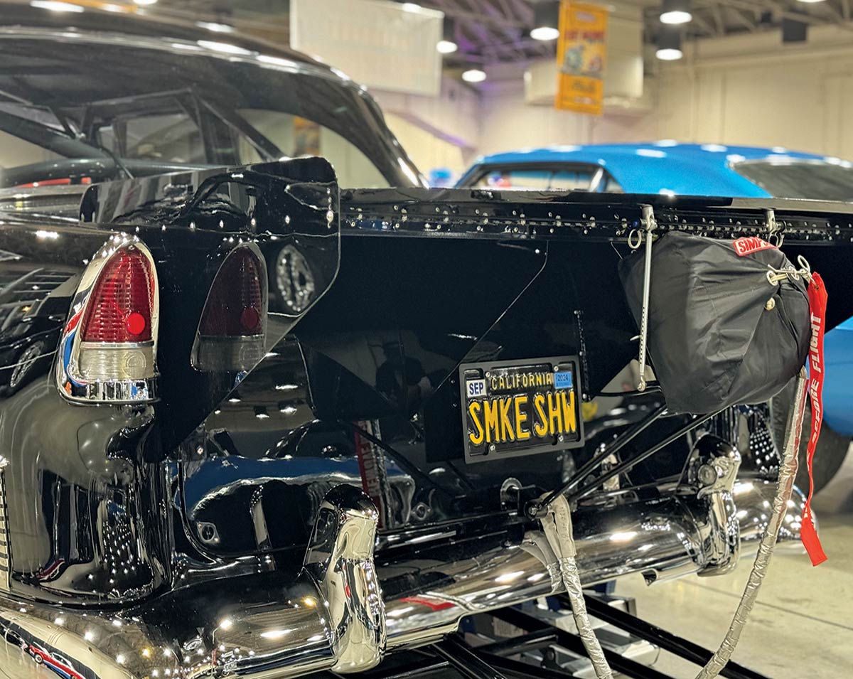 Landscape close-up angle rear photograph view of a vintage black Chevy car's trunk area as there is a bag equipped near the back chrome bumper inside The Grand National Roadster Show (GNRS) showroom venue