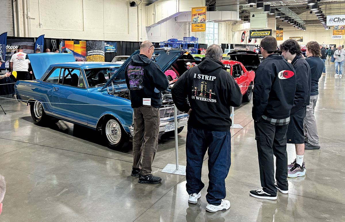 Landscape close-up angle photograph view of five individuals looking at a couple of vintage Chevy Camaro model cars (blue and red) displayed as they have their top front hoods open inside The Grand National Roadster Show (GNRS) showroom venue