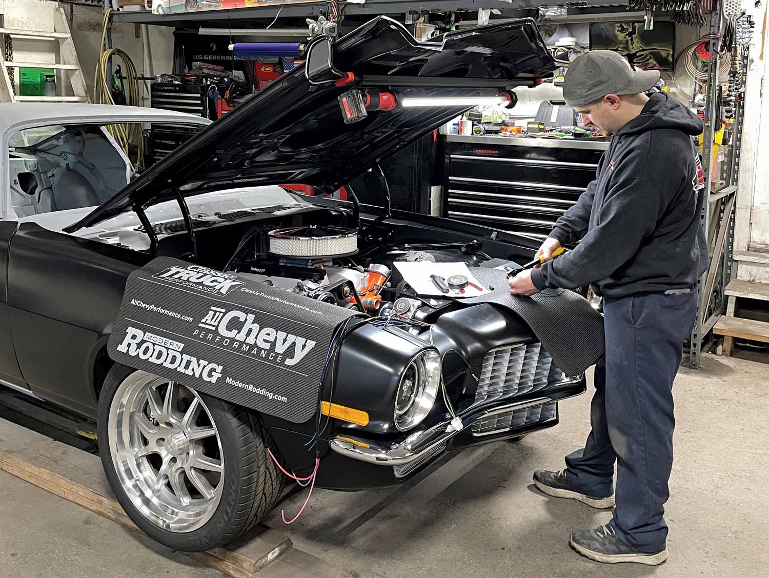 man standing in front of a black car with its hood open working on a car part