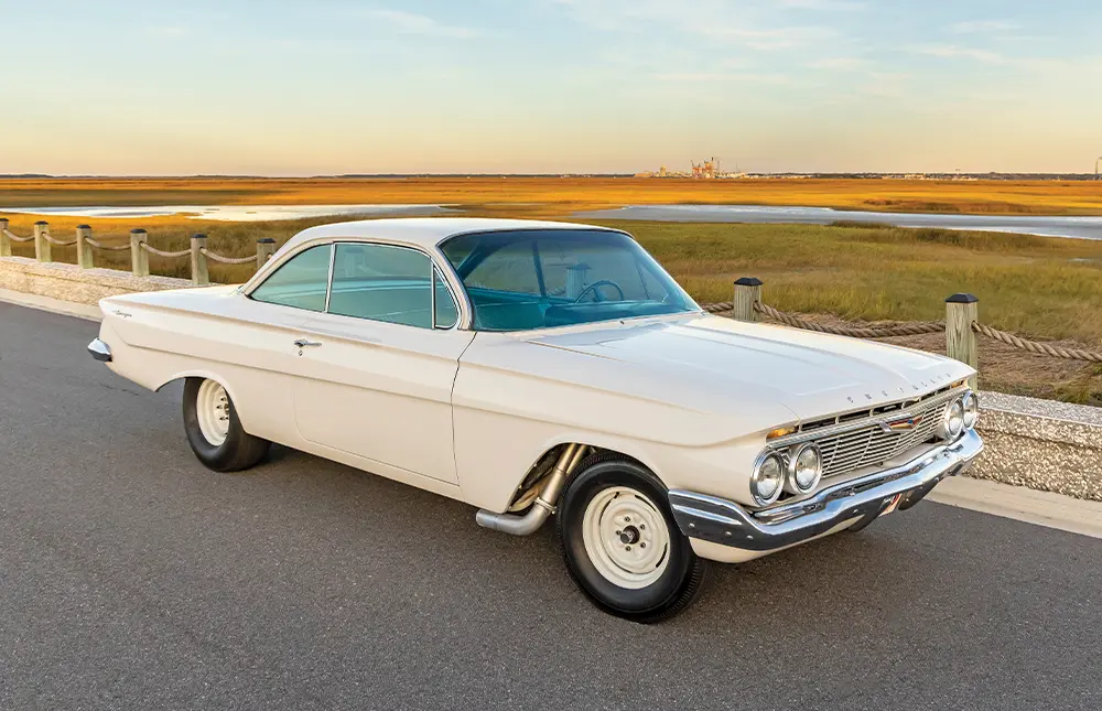 passenger side quarter view of the Ermine white ’61 Chevy Bel Air/Biscayne parked beside a rope and wood barrier on the edge of a vast marshy plain