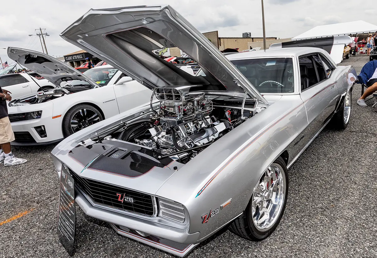 A highly modified silver 1969 Camaro Z28 with a supercharged engine, parked alongside a modern Camaro