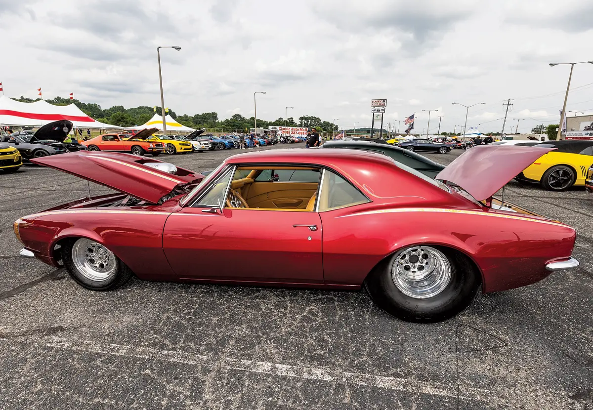 A side view of a highly modified red first-generation Camaro with drag racing slicks and a lowered stance, displayed outdoors