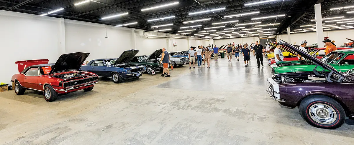 An indoor exhibition featuring multiple classic Camaros, with visitors walking between the rows of cars