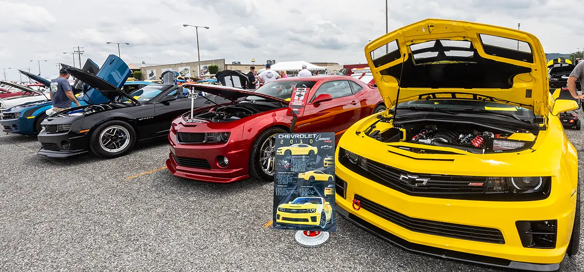 A lineup of different colored modern Camaros such as white, blue, black, red, yellow, etc. at a outdoor car show event in a parking lot setting area, with hoods up and engines on display as there are several visitors nearby these cars