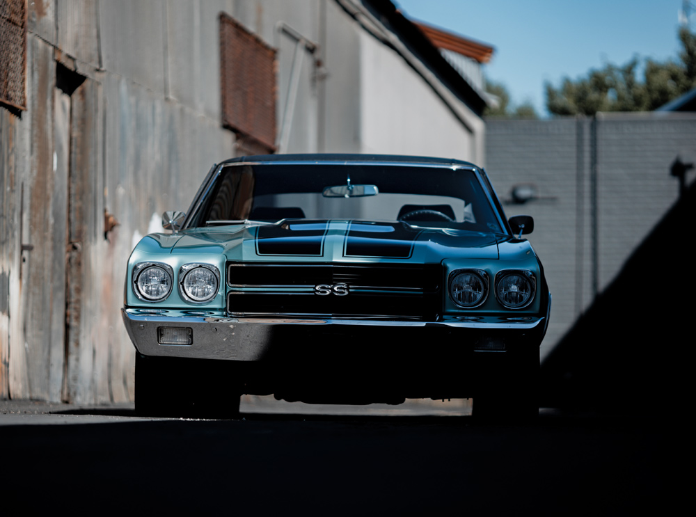 Front view of a blue classic muscle car with black racing stripes in an industrial alleyway.