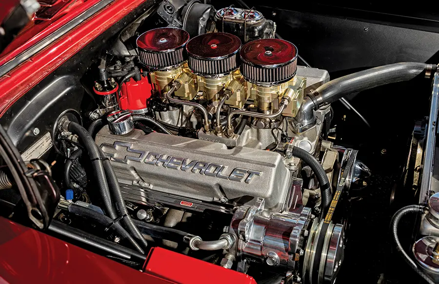 Close-up of a meticulously detailed Chevrolet V8 engine, featuring three chrome air filters over triple carburetors, with "CHEVROLET" embossed on the valve covers, installed in a red classic car.