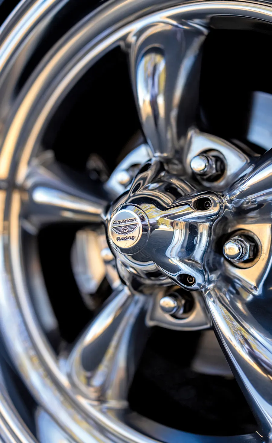 Detailed close-up of a highly polished chrome American Racing five-spoke wheel and center cap, showing lug nuts and reflections.
