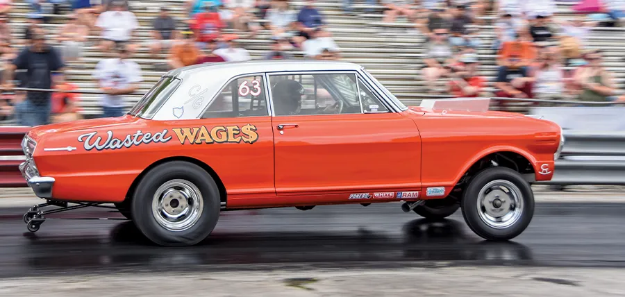 Orange and silver 'Wasted Wages' drag racing car with number 63, speeding down a track with blurred spectators in the background.