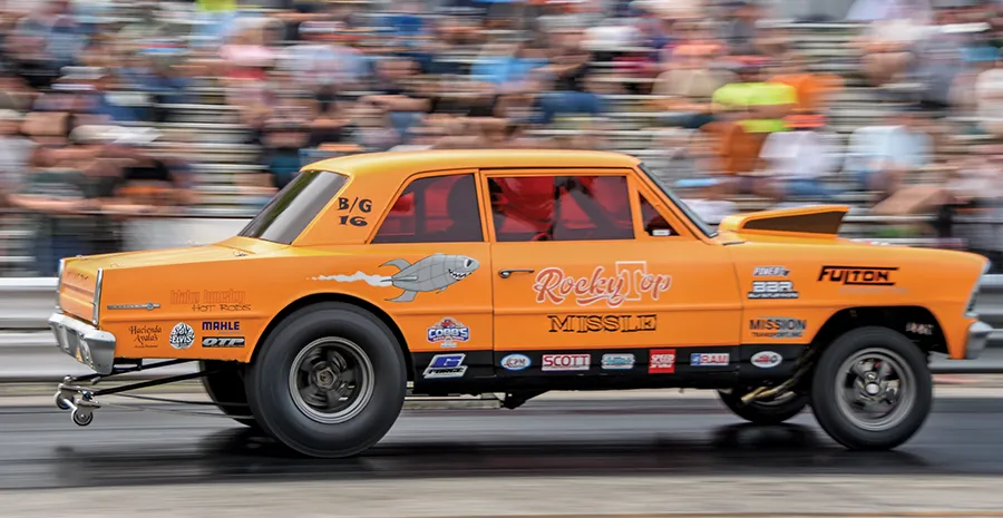 Side view of a vintage orange Chevrolet Nova gasser-style race car with a large fish graphic, speeding on a drag strip.