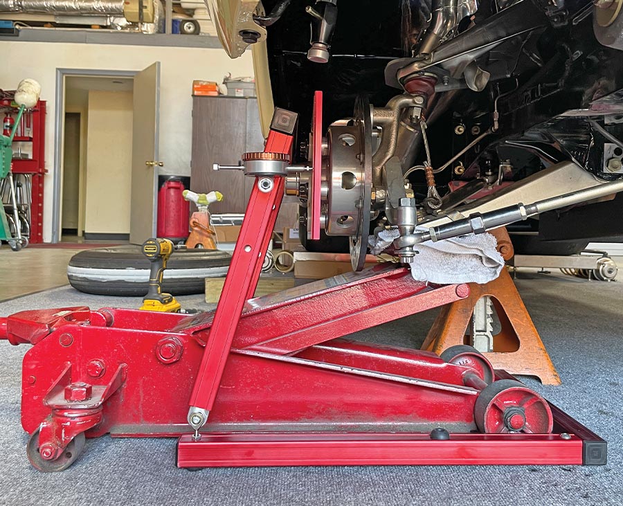 a close-up, low-angle shot shows a red hydraulic floor jack holding up the front suspension of a car in a garage