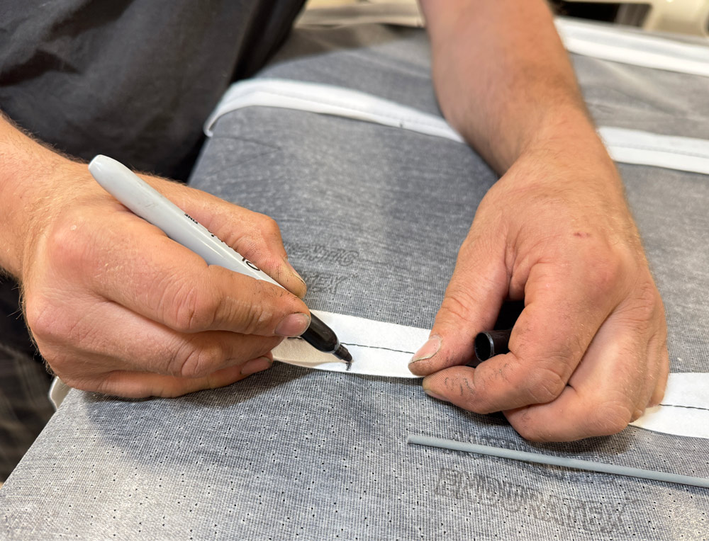 A close-up of hands marking a reference line with a black permanent marker on the white stitching of the headliner fabric.
