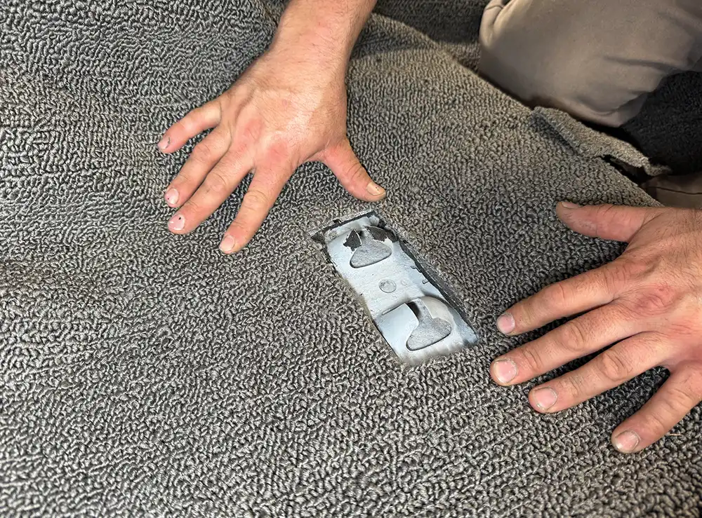 Hands pull the gray loop carpet back slightly to reveal an existing metal floor bracket for a seat or seatbelt attachment.