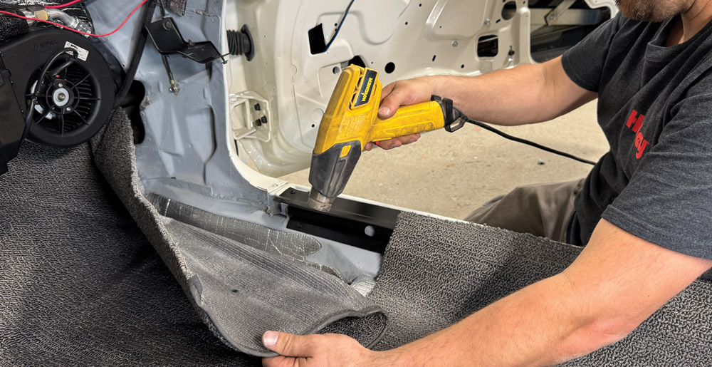 A person uses a yellow heat gun to warm the new gray carpet, making it more flexible for shaping and proper fitting.