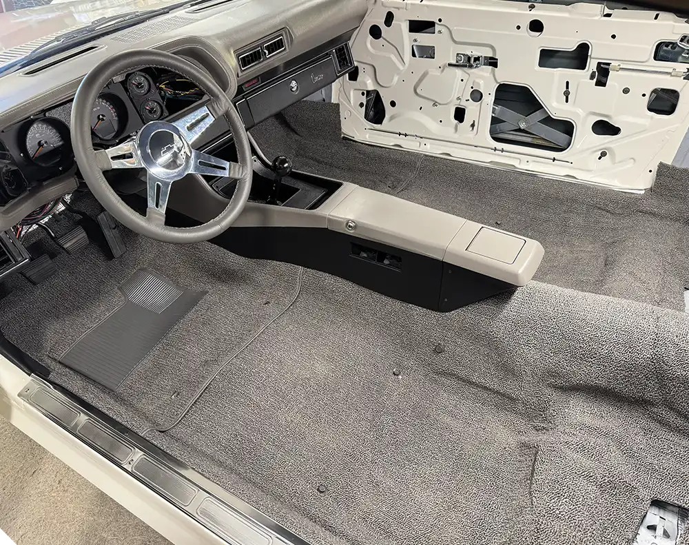 The car interior, now mostly complete, shows new gray carpet, a center console, steering wheel, and dashboard installed.