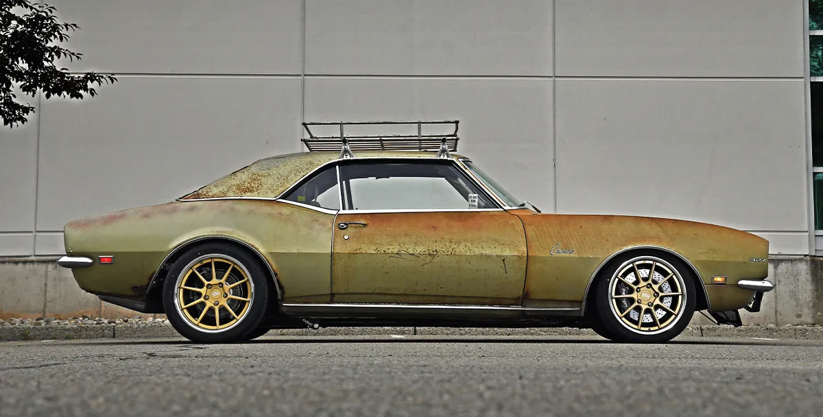 A side-view of a heavily patinaed olive-green 1967-69 Chevrolet Camaro coupe with a rusty roof rack, sporting gold aftermarket wheels with dark tires, parked on a street curb against a light gray wall.