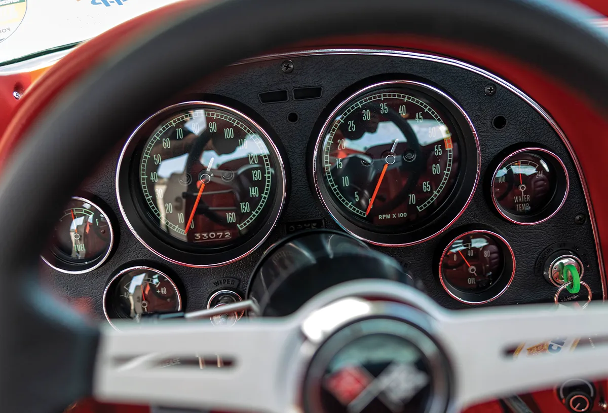 Close-up of the black dashboard and instrument cluster of a 1965 Corvette, showing the speedometer, tachometer, and auxiliary gauges through the steering wheel.