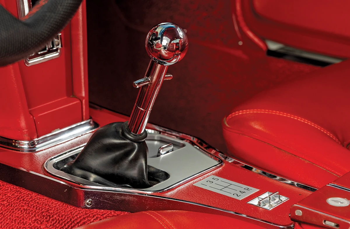 Close-up of a classic car interior showing a manual four-speed shifter with a chrome knob, set in a bright red carpet and red leather console.