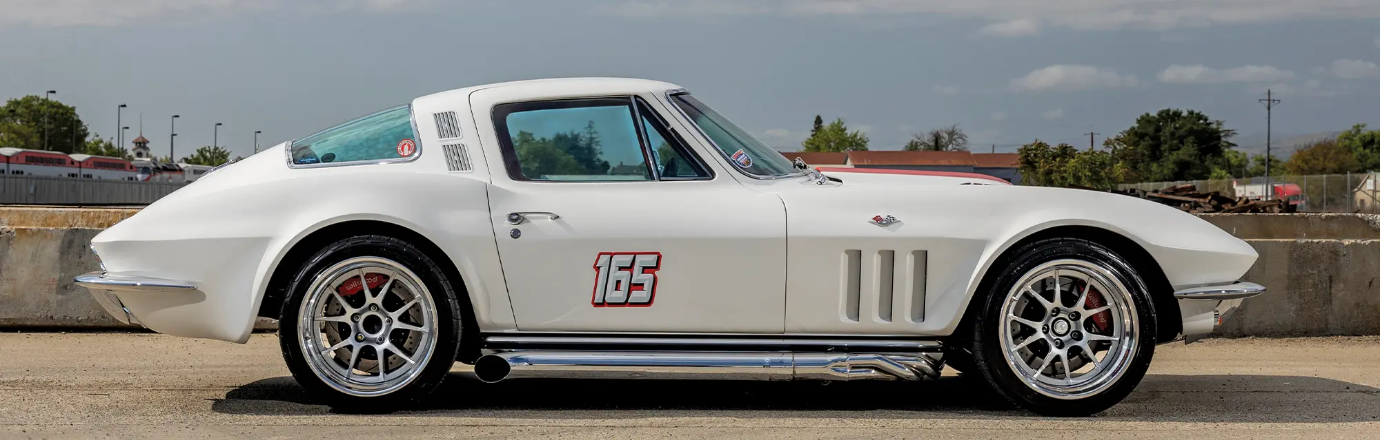 Profile view of a custom white 1965 Chevrolet Corvette with chrome side pipes, red racing numbers (165), and modern multi-spoke alloy wheels.