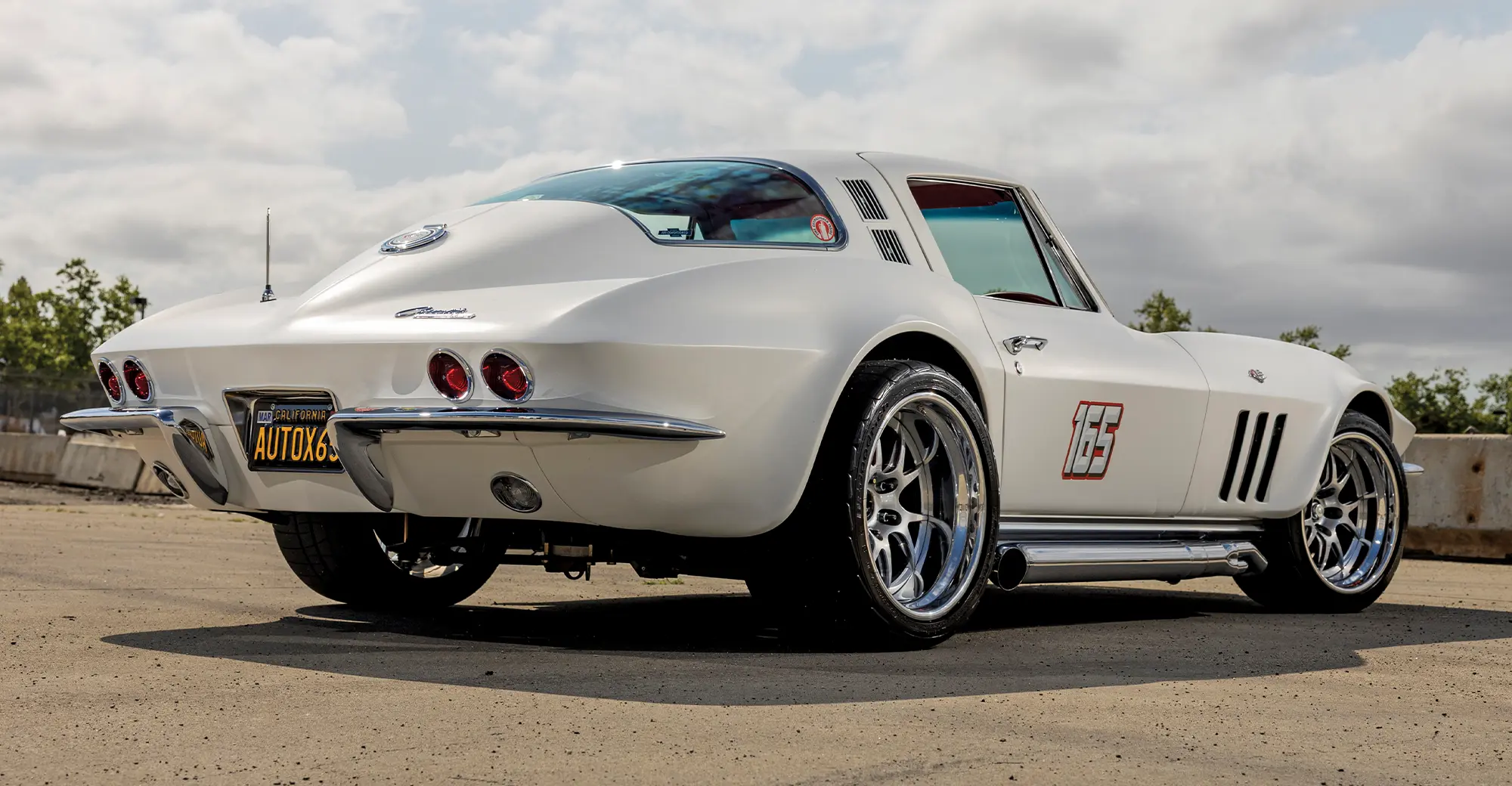 Rear three-quarter view of a custom white 1965 Chevrolet Corvette, showing the chrome side pipes, modern wheels, and distinctive rear end.