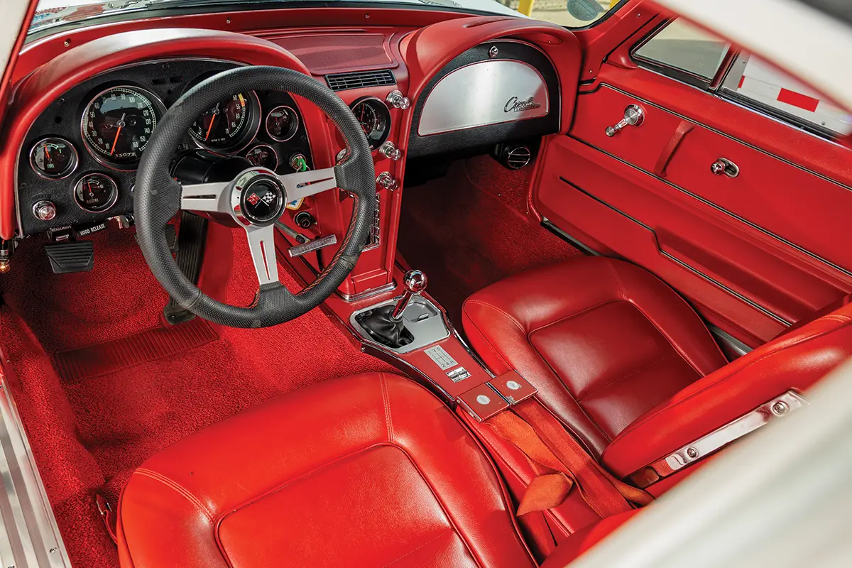 Interior view of a 1965 Chevrolet Corvette featuring custom vibrant red leather seats, red carpet, black dash, and a chrome manual shifter.