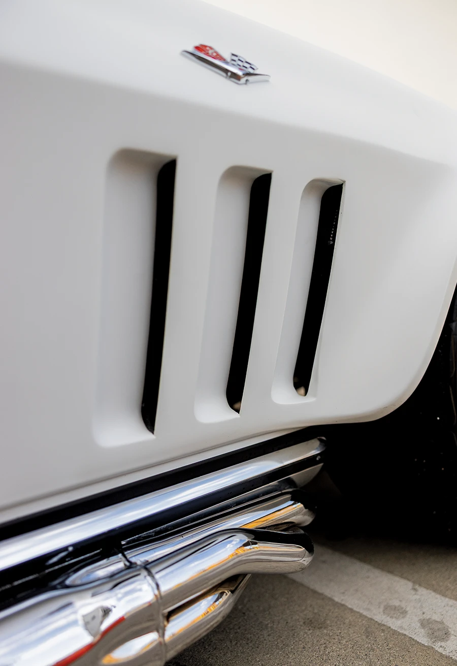 Close-up of the front fender of a white 1965 Corvette, highlighting the three vertical fender vents and the exposed chrome side pipe exhaust underneath.