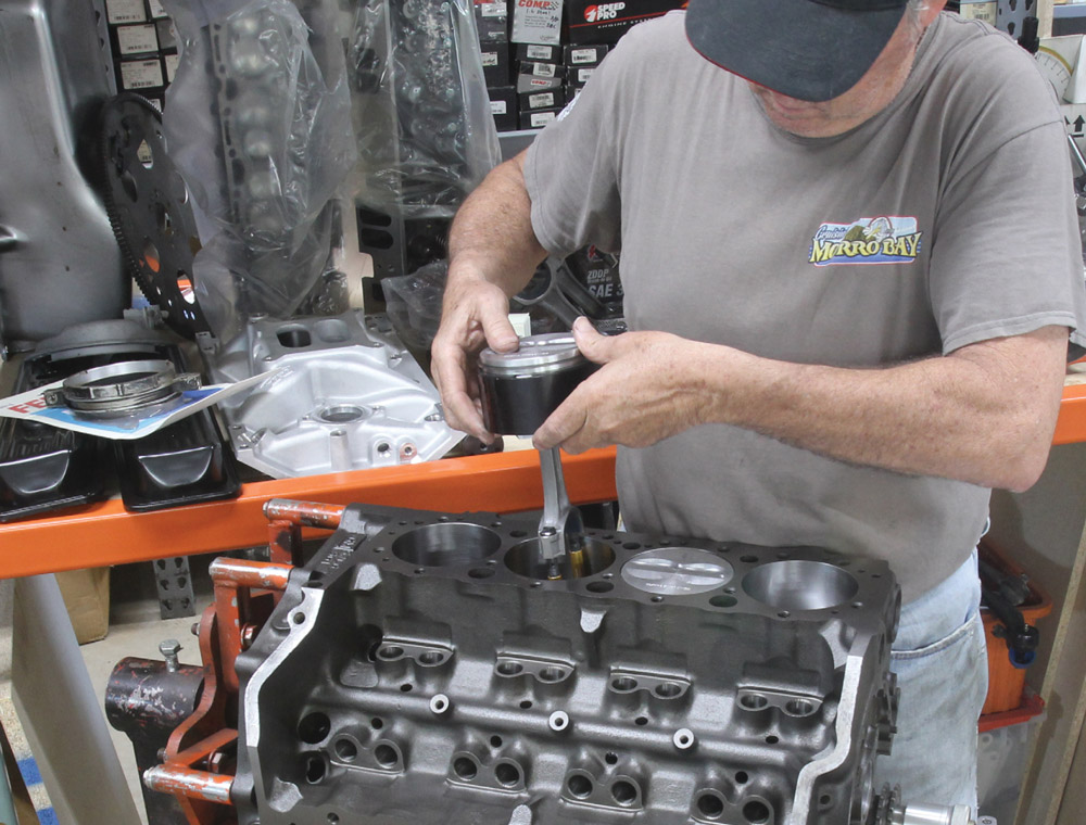 A mechanic installs a piston and connecting rod assembly into one of the cylinder bores of a bare Chevrolet small-block V8 engine block, using a ring compressor.