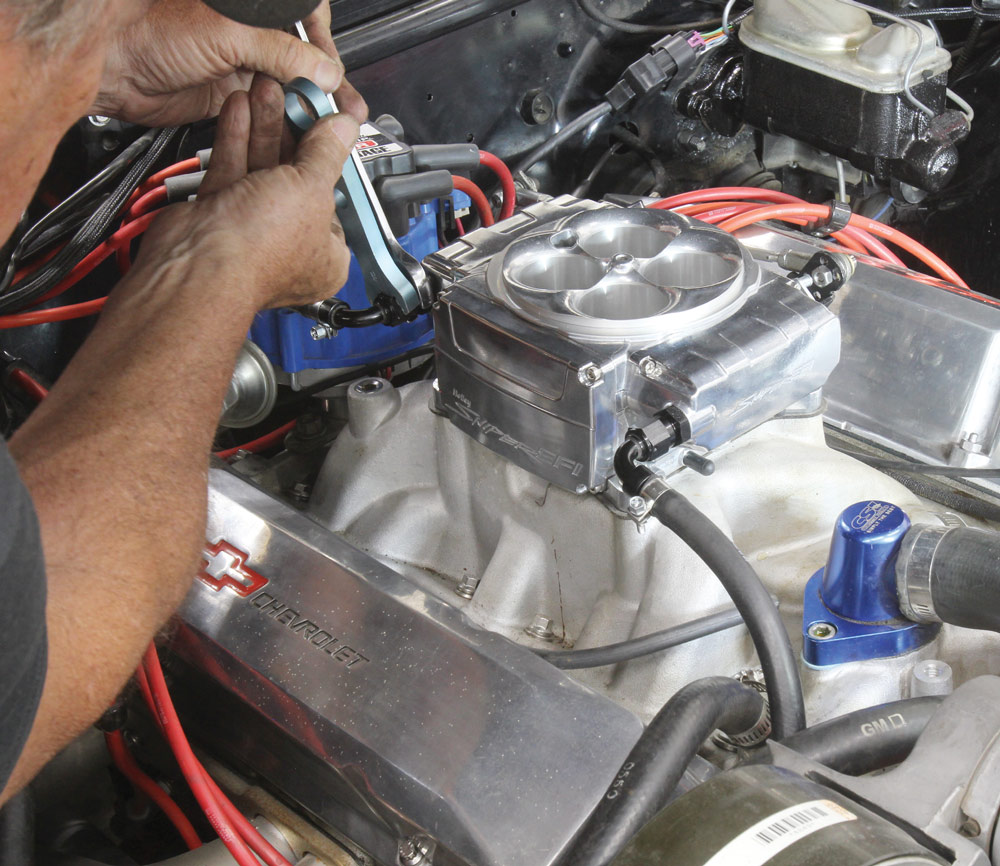 A mechanic is adjusting a highly polished, four-barrel throttle body, part of a Sniper EFI system, mounted on an aluminum intake manifold atop a Chevrolet V8 engine.