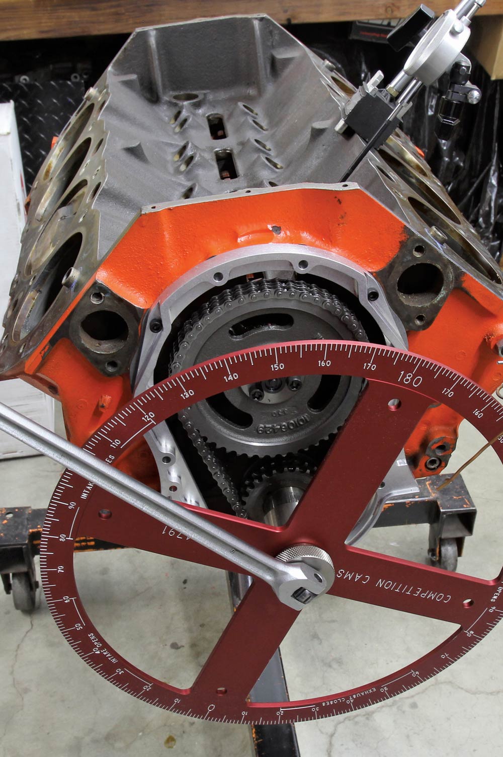 A large, red degree wheel is mounted to the crankshaft of an orange V8 engine block, positioned over the timing chain set. This tool is used to precisely check and adjust the engine's camshaft timing.