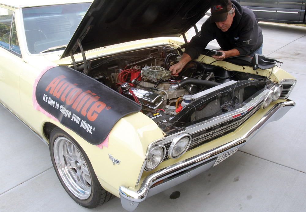 A man works on the engine of a yellow 1966 Chevelle with the hood open. The car is protected by an Autolite fender cover. He is adjusting the carburetor or ignition while holding a mobile device.