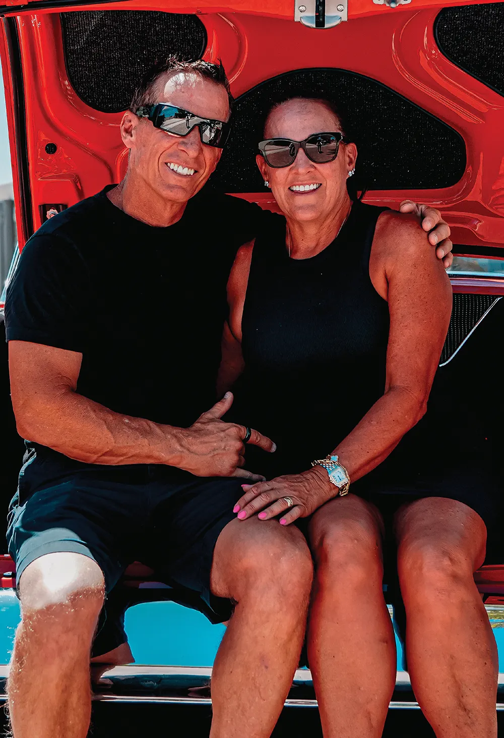 Close-up portrait of a smiling man and woman wearing sunglasses, sitting together on the trunk ledge of the orange 1956 Chevrolet 210 sedan.