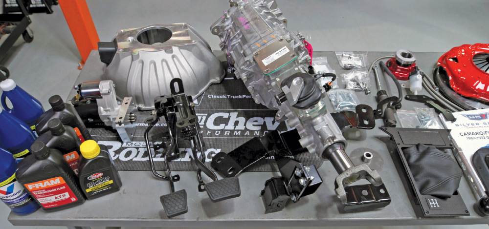Overhead view of a manual transmission kit, including the transmission, bellhousing, clutch pedals, fluids, and small hardware components laid out on a table.