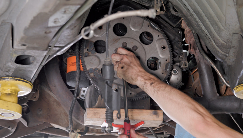 A mechanic's hand works underneath a raised vehicle, using a wrench to remove bolts from the flexplate near the engine oil pan.