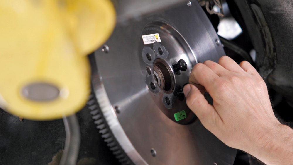 Close-up of a mechanic's hand installing a new metallic flywheel onto the rear of an engine, with the pilot bearing visible in the center.