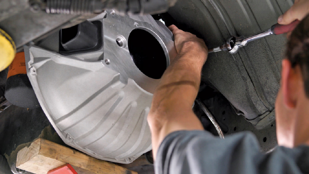 A mechanic's view from beneath a raised car as he bolts the new silver bellhousing onto the rear of the engine block.